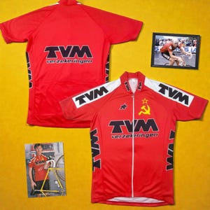 May include: Red cycling jersey with white accents, featuring the "TVM verzekeringen" logo in black. The jersey has a full zipper and a star and hammer and sickle emblem. A framed photo of a cyclist is also visible.
