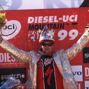 May include: A person wearing a sequined jacket, crown, and sunglasses, holding a bouquet. The background features a red banner with "DIESEL-UCI MOUNTAIN BIKE 99" text and logos. The person is celebrating a victory.