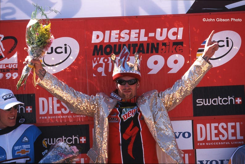 May include: A person wearing a sequined jacket, crown, and sunglasses, holding a bouquet. The background features a red banner with "DIESEL-UCI MOUNTAIN BIKE 99" text and logos. The person is celebrating a victory.