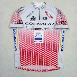 May include: A white and red short-sleeved cycling jersey featuring a honeycomb pattern. The front includes a half-zip collar and multiple printed logos such as Colnago, Landbouwkrediet, Olympus, and Esprinet, alongside various black text and graphic elements.