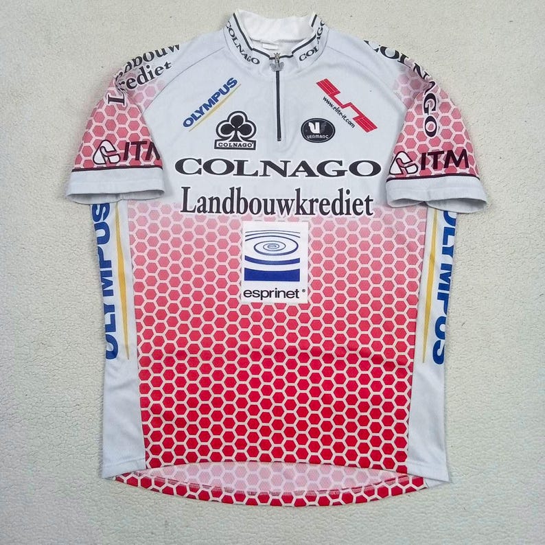May include: A white and red short-sleeved cycling jersey featuring a honeycomb pattern. The front includes a half-zip collar and multiple printed logos such as Colnago, Landbouwkrediet, Olympus, and Esprinet, alongside various black text and graphic elements.