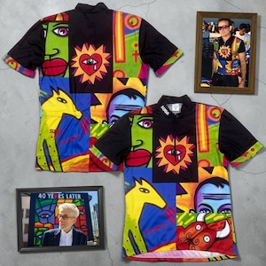 Puede incluir: Dos camisas de manga corta con un diseño abstracto y colorido que presenta caras, un corazón con un ojo y un caballo amarillo. Las camisas son negras con la palabra "SUGA" en el cuello. Dos fotos enmarcadas también están en la imagen.