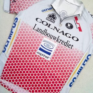 May include: A white cycling jersey with a red honeycomb gradient pattern on the front. The shirt features a short zip at the collar and various printed logos including Colnago, Landbouwkrediet, Esprinet, and Olympus in blue, black, and red text.