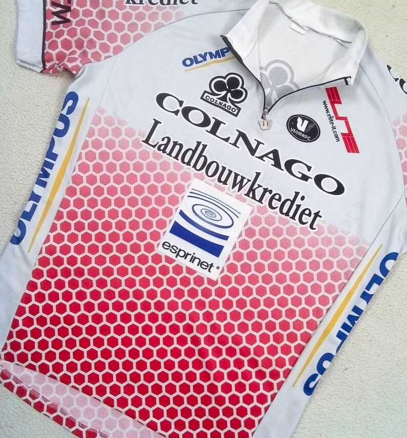 May include: A white cycling jersey with a red honeycomb gradient pattern on the front. The shirt features a short zip at the collar and various printed logos including Colnago, Landbouwkrediet, Esprinet, and Olympus in blue, black, and red text.