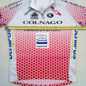 May include: A white and pink cycling jersey with a honeycomb pattern, featuring logos for Colnago, Landbouwkrediet, Olympus, and Esprinet. A yellow measuring tape is placed across the chest, showing a width of approximately 58.5 cm.