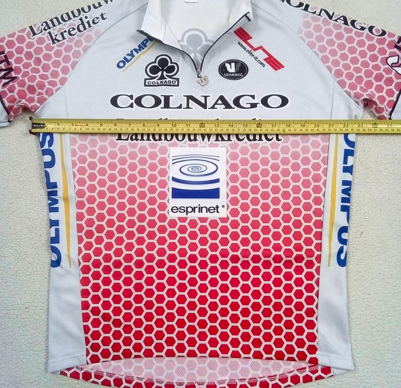 May include: A white and pink cycling jersey with a honeycomb pattern, featuring logos for Colnago, Landbouwkrediet, Olympus, and Esprinet. A yellow measuring tape is placed across the chest, showing a width of approximately 58.5 cm.