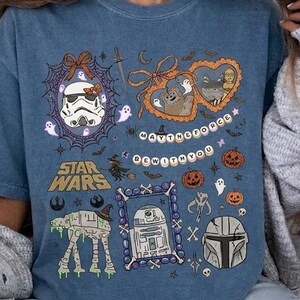 May include: A blue short-sleeve t-shirt featuring a Star Wars Halloween graphic print. The design includes a Stormtrooper, R2-D2, an AT-AT, and a Mandalorian helmet surrounded by pumpkins, ghosts, and the text MAY THE FORCE BE WITH YOU.