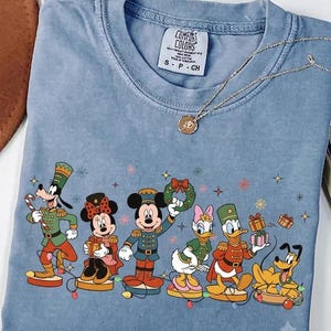 May include: A blue short-sleeved t-shirt featuring a graphic print of Disney characters dressed as nutcrackers, including Goofy, Minnie Mouse, Mickey Mouse, Daisy Duck, Donald Duck, and Pluto, with the Comfort Colors brand label visible.
