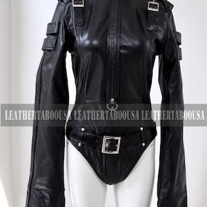 May include: A black leather bodysuit featuring a high collar with a metal ring, long sleeves with buckled straps, and a waist belt with a silver buckle. The garment includes shoulder epaulets and the text LEATHERTABOOUSA printed across the chest.