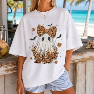 May include: A white short-sleeved t-shirt featuring a central graphic of a ghost wearing a large leopard-print bow. The ghost has a leopard-print pattern at the base, surrounded by small black bats, orange autumn leaves, and tiny star accents.