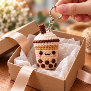 May include: A crocheted boba tea keyring featuring a tan cup with dark brown tapioca pearls, a cream-coloured top, and a brown straw. The keyring has a metal ring and chain attached to the top, showcasing a small, smiling face with pink cheeks.