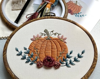 May include: An embroidered pumpkin design in an oval wooden hoop. The pumpkin is orange with green vines and surrounded by blue-green leaves and burgundy flowers. A pen, embroidery floss, and a pattern are in the background.