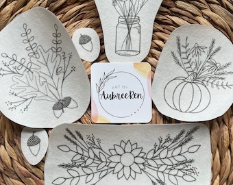 May include: A collection of fabric patches with botanical designs, including a sunflower, a bouquet in a jar, and fall leaves. The patches are outlined in black on a light background. A small card with the text "ART BY Aubree Ren" is also present.