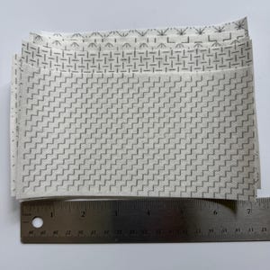 May include: A stack of rectangular white fabric swatches featuring various gray geometric patterns, including zigzags, dashes, and starbursts. A metal ruler is positioned along the bottom edge, showing the width is approximately 7 inches.