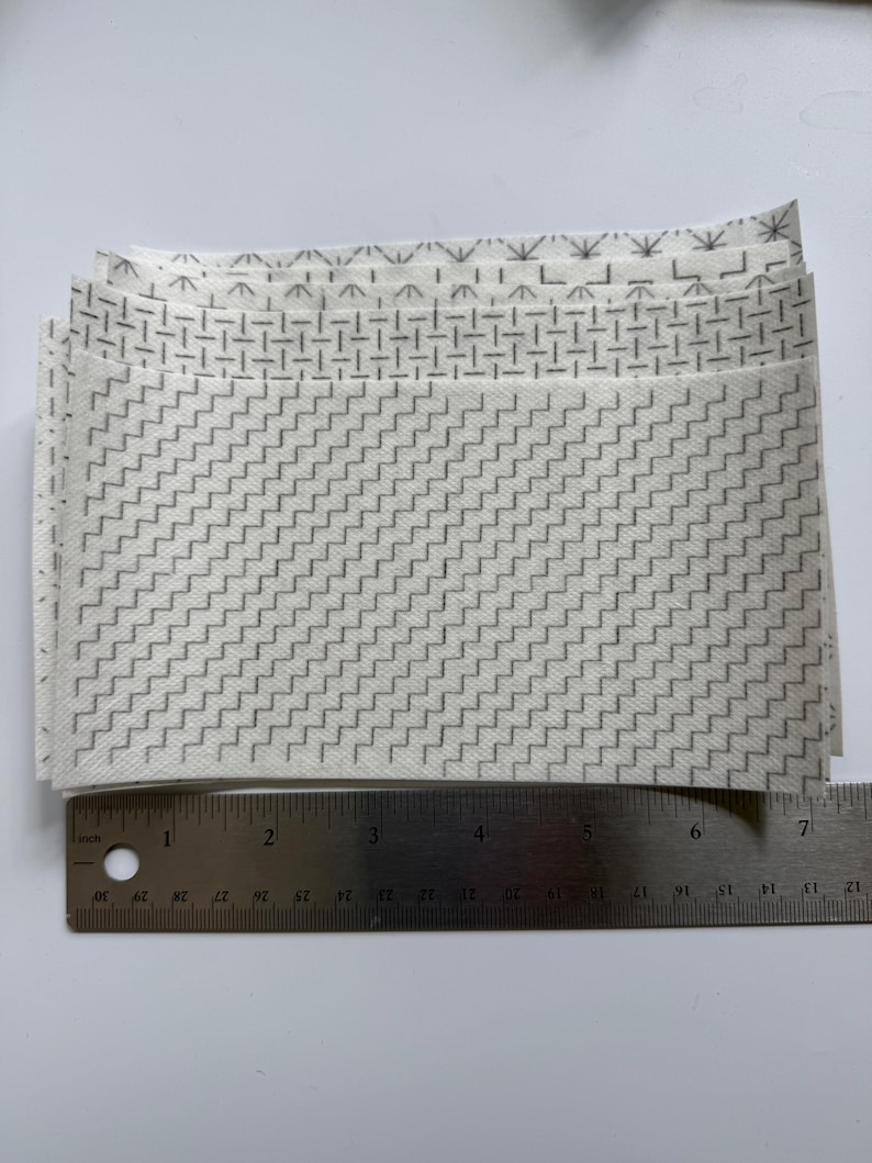 May include: A stack of rectangular white fabric swatches featuring various gray geometric patterns, including zigzags, dashes, and starbursts. A metal ruler is positioned along the bottom edge, showing the width is approximately 7 inches.