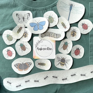 May include: A dark green t-shirt featuring various insect-themed fabric patches, including butterflies, beetles, a caterpillar, a dragonfly, a bee, and a line of ants. A square white card with the text Art by AubreeRen is centered among them.