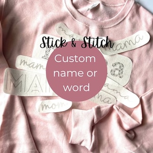 May include: A pink sweatshirt with the words "Stick & Stitch" and "Custom name or word" printed on it. There are several white iron-on letters on the sweatshirt, spelling out the word "mama".
