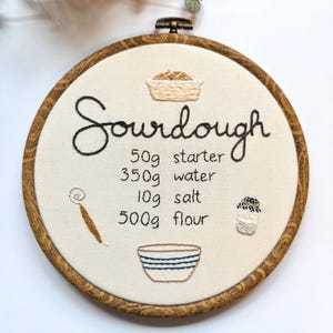 May include: Embroidered sourdough recipe on a round, wooden hoop. The text "Sourdough" is in cursive, with ingredient amounts listed below: 50g starter, 350g water, 10g salt, and 500g flour. Illustrations of a loaf, bowl, and jar are included.