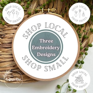 May include: A wooden embroidery hoop with a white fabric design. The design features the text "SHOP LOCAL" and "SHOP SMALL" around a central design that says "Three Embroidery Designs". Additional small circular designs with botanical illustrations are also present.