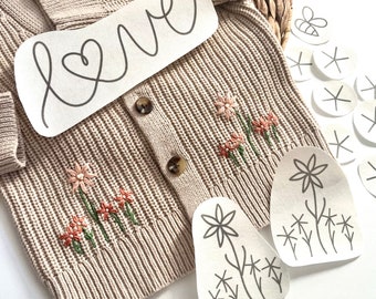 May include: A beige knitted cardigan with embroidered flowers and the word "love" written on a piece of paper. There are also several paper cutouts of flowers and a bee.