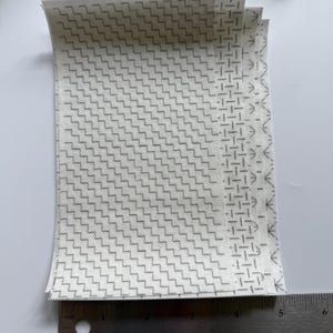 May include: A stack of rectangular white fabric sheets featuring a repeating gray geometric pattern of zigzags and lines. A metal ruler is positioned at the bottom, showing the width of the sheets is approximately 5.25 inches wide.