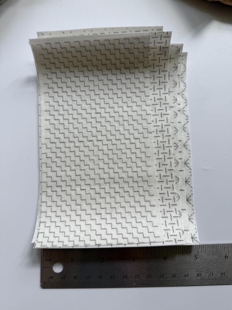 May include: A stack of rectangular white fabric sheets featuring a repeating gray geometric pattern of zigzags and lines. A metal ruler is positioned at the bottom, showing the width of the sheets is approximately 5.25 inches wide.
