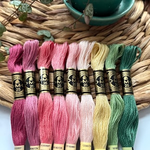 May include: Eight spools of embroidery floss in shades of pink, peach, cream, yellow, and green. Each spool is labeled "DMC" and "M-25" with a number.