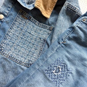 May include: A light blue denim jacket featuring a tan corduroy collar and a chest pocket adorned with white star-like embroidery. The sleeve displays a blue geometric embroidered pattern surrounding a small distressed hole in the fabric.