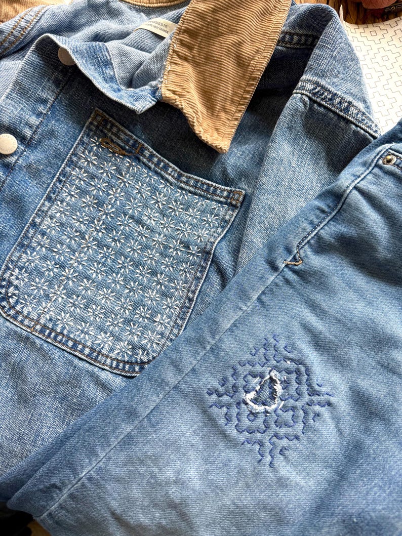 May include: A light blue denim jacket featuring a tan corduroy collar and a chest pocket adorned with white star-like embroidery. The sleeve displays a blue geometric embroidered pattern surrounding a small distressed hole in the fabric.
