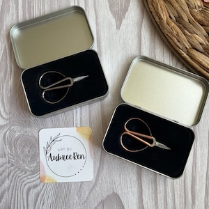 May include: Two small metal scissors with rounded handles, one silver and one copper-toned, each resting in a rectangular silver tin with a black foam insert. A square white card with a circular logo and the text Art By Aubree Ren is included.