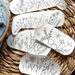 Op de afbeelding: Vijf witte stoffen patches met zwarte borduursels van verschillende bloemenmotieven. De patches zijn gestapeld, met de grootste patch onderaan en de kleinste patch bovenaan.