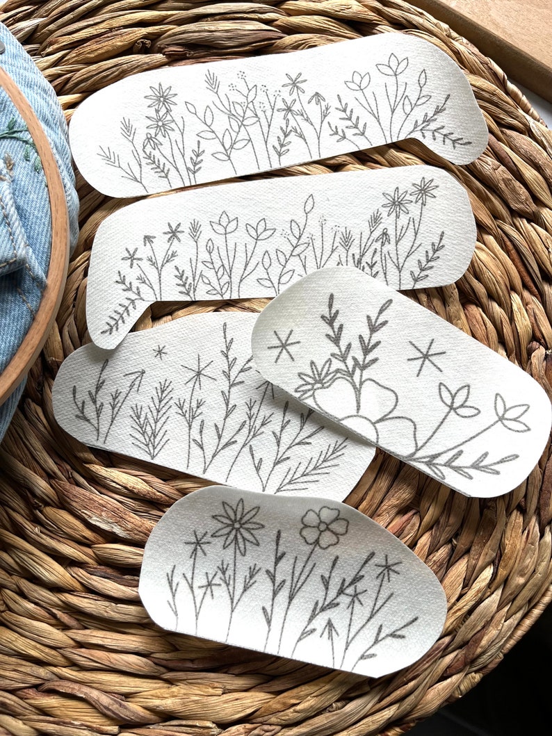 Op de afbeelding: Vijf witte stoffen patches met zwarte borduursels van verschillende bloemenmotieven. De patches zijn gestapeld, met de grootste patch onderaan en de kleinste patch bovenaan.