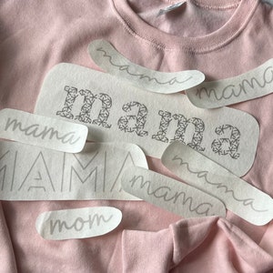 May include: A pink sweatshirt with several white iron-on vinyl designs that say "mama" in different fonts and styles.