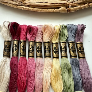 May include: A selection of DMC embroidery floss in shades of cream, burgundy, pink, peach, yellow, green, grey, and purple. Each skein is labelled with "DMC 25" and measures 8 m (8.7 yd).