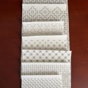 May include: A vertical stack of seven rectangular fabric swatches, each featuring a distinct white background with a repeating gray geometric pattern. The designs include stepped squares, radiating lines, crosses, starbursts, and woven grid motifs.