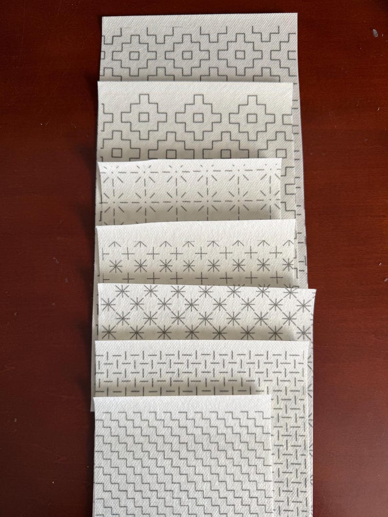 May include: A vertical stack of seven rectangular fabric swatches, each featuring a distinct white background with a repeating gray geometric pattern. The designs include stepped squares, radiating lines, crosses, starbursts, and woven grid motifs.