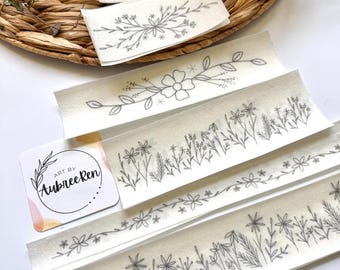 Op de afbeelding: Een verzameling bloemendesignstickers met fijne grijze lijntekeningen. De stickers tonen verschillende botanische illustraties, waaronder bloemen, bladeren en sprietjes. Een kleine kaart met de tekst "ART BY AubreeRen" is ook aanwezig. De stickers zijn gerangschikt op een geweven oppervlak.