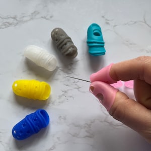 May include: Assortment of colorful finger thimbles made of silicone, designed to protect fingers while sewing. The thimbles come in blue, pink, yellow, white, and gray. A sewing needle is also visible in the image.