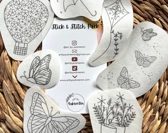 May include: A set of 8 iron-on embroidery patches featuring various designs, including a hot air balloon, umbrella, kite, butterflies, flowers, and rain boots. The patches are white with black outlines and are arranged on a woven wicker surface. The text "Stick & Stitch Pack" is visible in the center of the image.