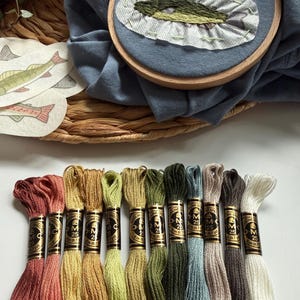 以下が含まれることがあります： 木製の刺繍枠に青い布と緑色の魚の刺繍デザインが施されており、魚の形をした布の切り抜きと、赤、金、緑、青、白の色合いの12本のDMC刺繍糸の束が並べられています。