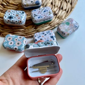 May include: A small pink metal box with a floral design and a hinged lid. The box is open and contains a small sewing kit with needles, thread, and a thimble.