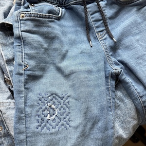 May include: Light blue denim jeans featuring an elastic waistband with a black drawstring. The front of the left leg displays a square patch of intricate dark blue geometric embroidery with a small frayed hole in the center of the stitched design.