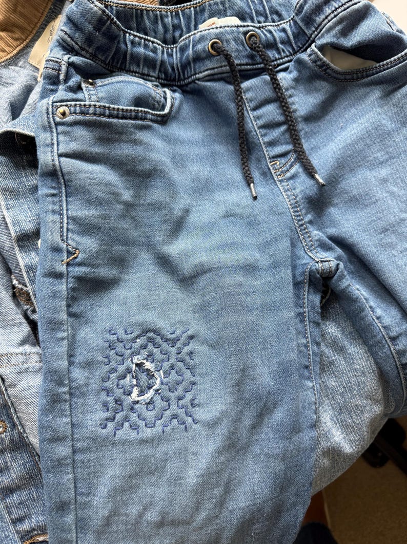 May include: Light blue denim jeans featuring an elastic waistband with a black drawstring. The front of the left leg displays a square patch of intricate dark blue geometric embroidery with a small frayed hole in the center of the stitched design.