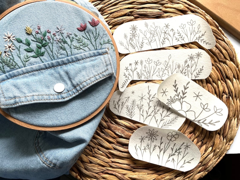 Op de afbeelding: Vijf borduurpatronen voor bloemmotieven op een geweven rieten achtergrond. De patronen zijn gedrukt op wit papier en bevatten verschillende bloemmotieven, waaronder bladeren, stengels en bloemen.