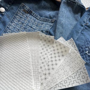 May include: A collection of white rectangular sheets featuring various gray geometric patterns, including zigzags, grids, stars, and stepped lines, fanned out over a blue denim jacket with a brown corduroy collar and white stitched details.