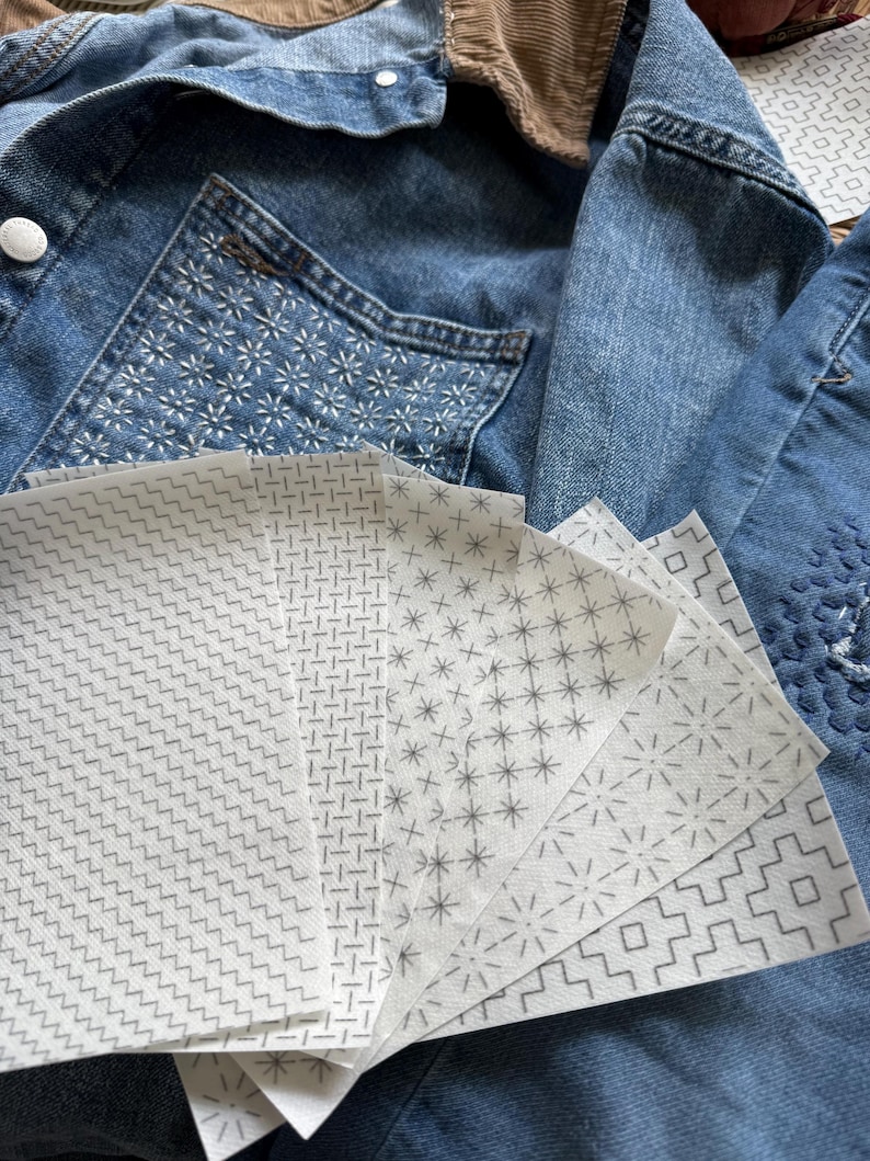 May include: A collection of white rectangular sheets featuring various gray geometric patterns, including zigzags, grids, stars, and stepped lines, fanned out over a blue denim jacket with a brown corduroy collar and white stitched details.