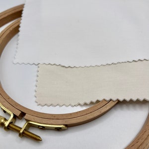 May include: Two wooden embroidery hoops with a white and a cream fabric stretched over them. The hoops are nested together and the fabric is slightly wrinkled.