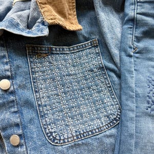 May include: A blue denim jacket featuring a tan corduroy collar, silver-toned metal buttons, and a chest pocket adorned with a white stitched floral pattern. The fabric has a light wash finish, and the interior label reads Oversized Fit.