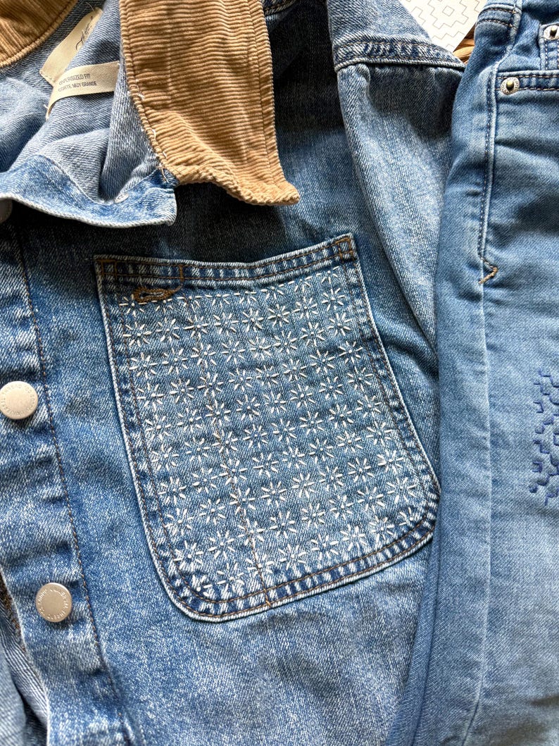 May include: A blue denim jacket featuring a tan corduroy collar, silver-toned metal buttons, and a chest pocket adorned with a white stitched floral pattern. The fabric has a light wash finish, and the interior label reads Oversized Fit.