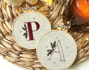 May include: Two circular wooden embroidery hoops containing beige fabric with hand-stitched letters P and A. The letter P is deep red, and the letter A is outlined in red and white thread. Both feature green pine sprig accents and small star motifs.